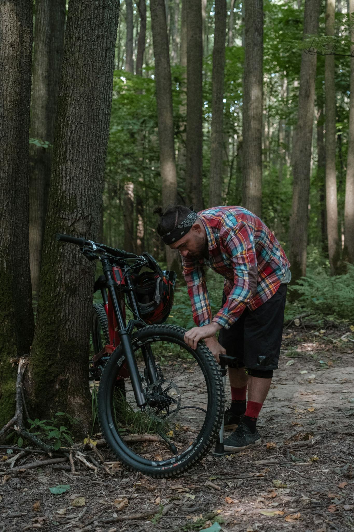 Man examines his mountain bike tire during a forest adventure, surrounded by tall trees.